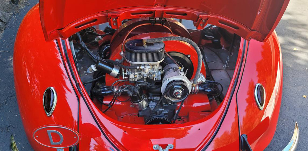 A detailed top-down view of the clean air-cooled engine bay of a red VW Beetle shorty. The engine features a single carburetor with a rectangular air cleaner and black components set against color-matched red tinware.