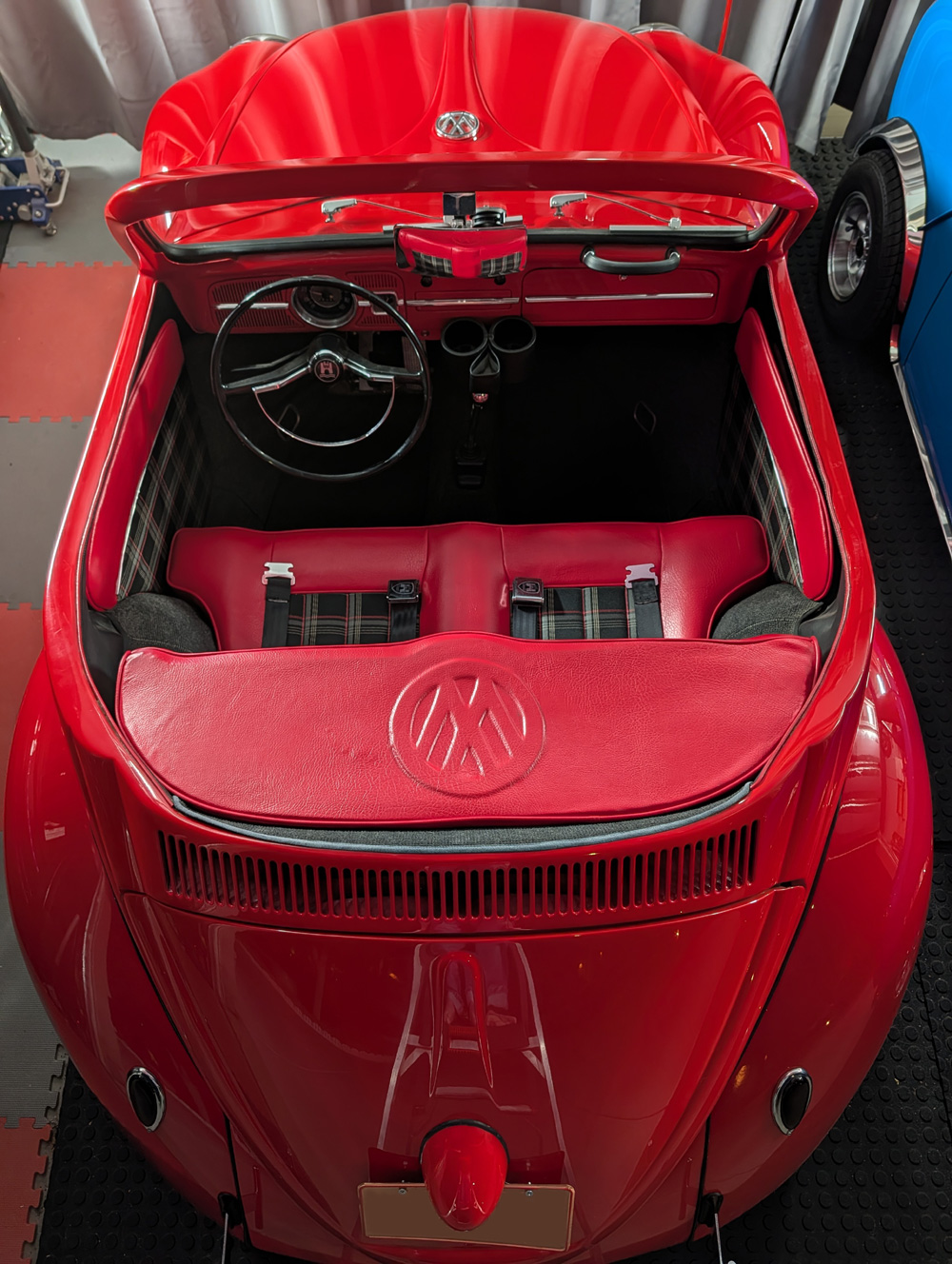 Interior view of a custom red VW Beetle shorty convertible showing a black two-spoke steering wheel, a body-matched red dashboard with a classic speedometer, and grey vinyl seats with red piping.