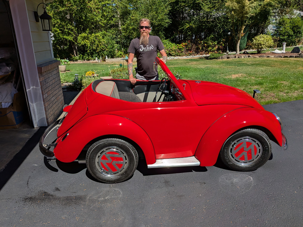 Owner Robert Basil standing behind his custom red Volkswagen Beetle shorty convertible on a paved driveway, showcasing the significantly shortened wheelbase and custom red VW logo wheels.