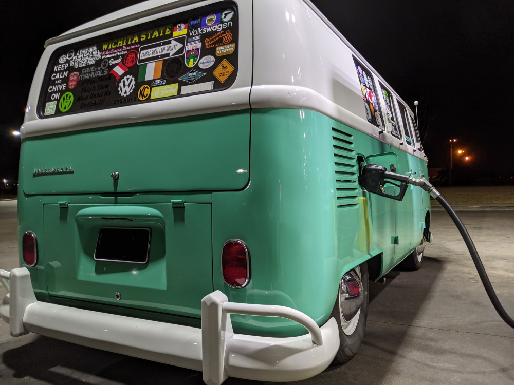 Rear view of a turquoise and white VW Bus parked on grass in a wooded area.