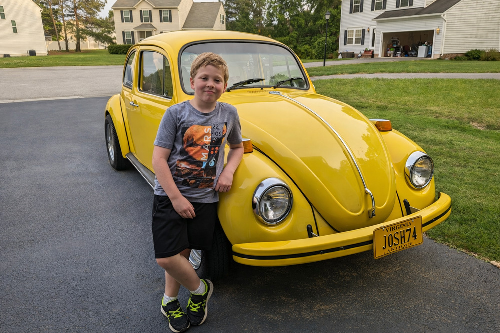 Josh standing beside a yellow Volkswagen Beetle with JOSH74 license plate in a suburban driveway