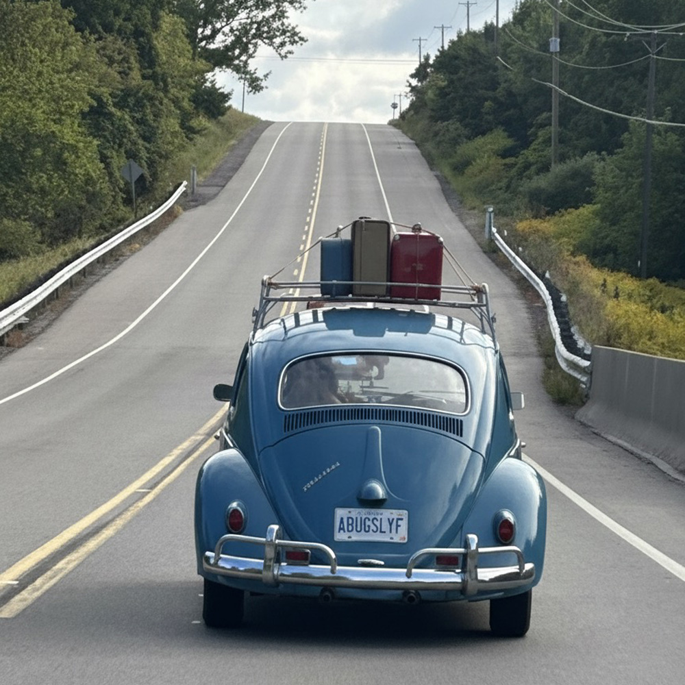 Blue Volkswagen Beetle driving uphill with roof-rack suitcases on two-lane road