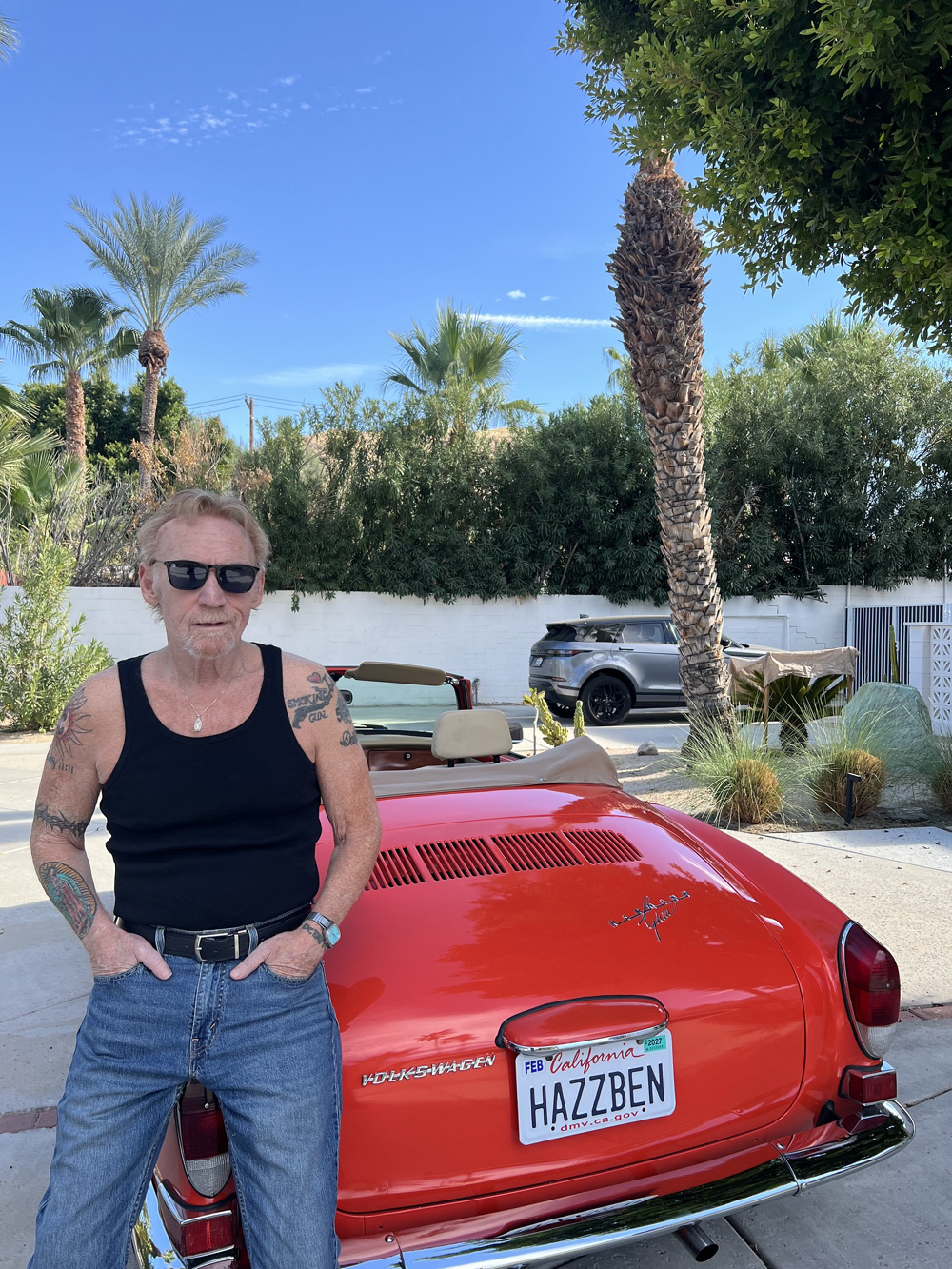 Danny Bonaduce posing with his red Karmann Ghia convertible in a sunny Palm Springs setting with palm trees behind him