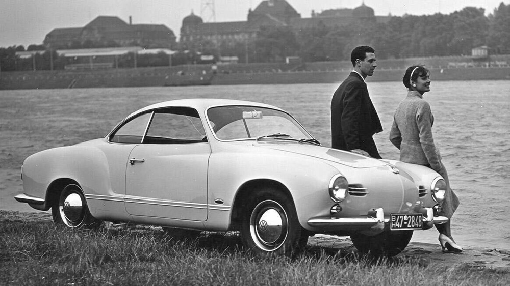 Vintage black and white photo of a classic Karmann Ghia coupe parked by the water with a stylish couple