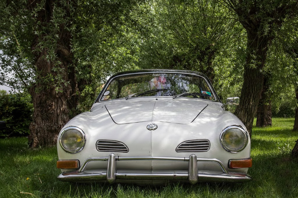 Classic white Karmann Ghia coupe parked on green grass between large trees