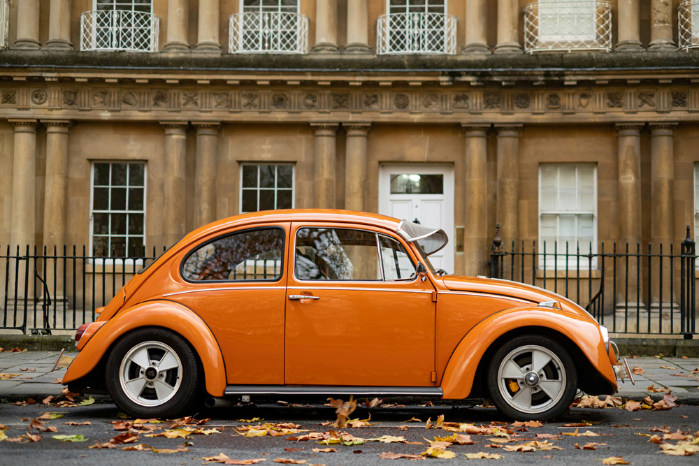 Orange vintage Volkswagen Beetle parked on a street in front of a neoclassical building with tall columns and autumn leaves on the ground.