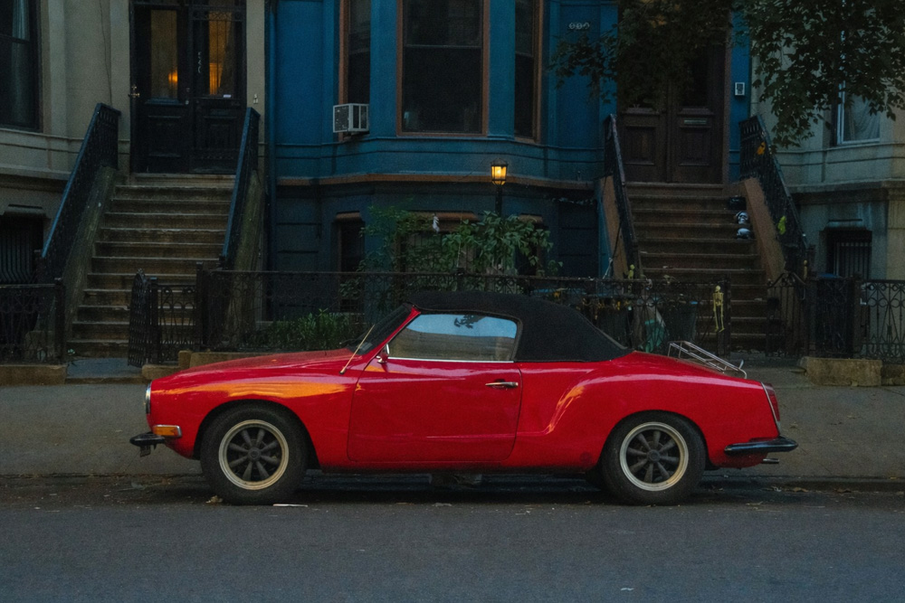 Red Karmann Ghia convertible parked on a residential street in front of brownstone buildings with stoops and a lit streetlamp.