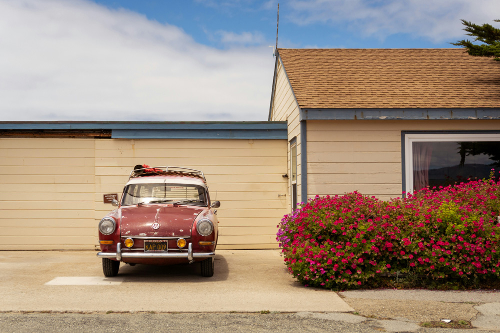 Red vintage Volkswagen Squareback parked in front of a wooden garage and a house with blooming pink flowers.