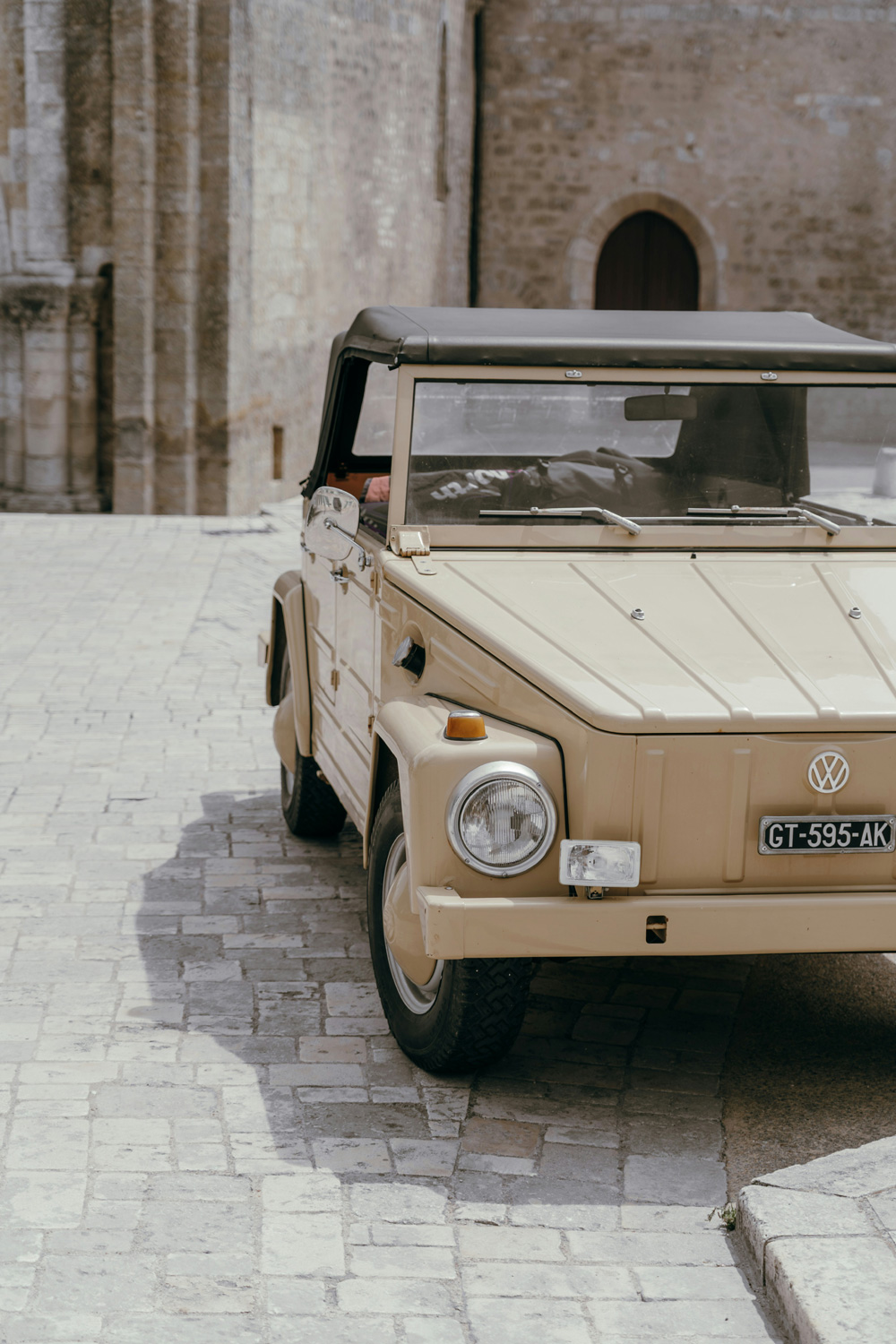 Beige vintage Volkswagen Thing parked on a cobblestone street in front of a stone building with an arched wooden door.