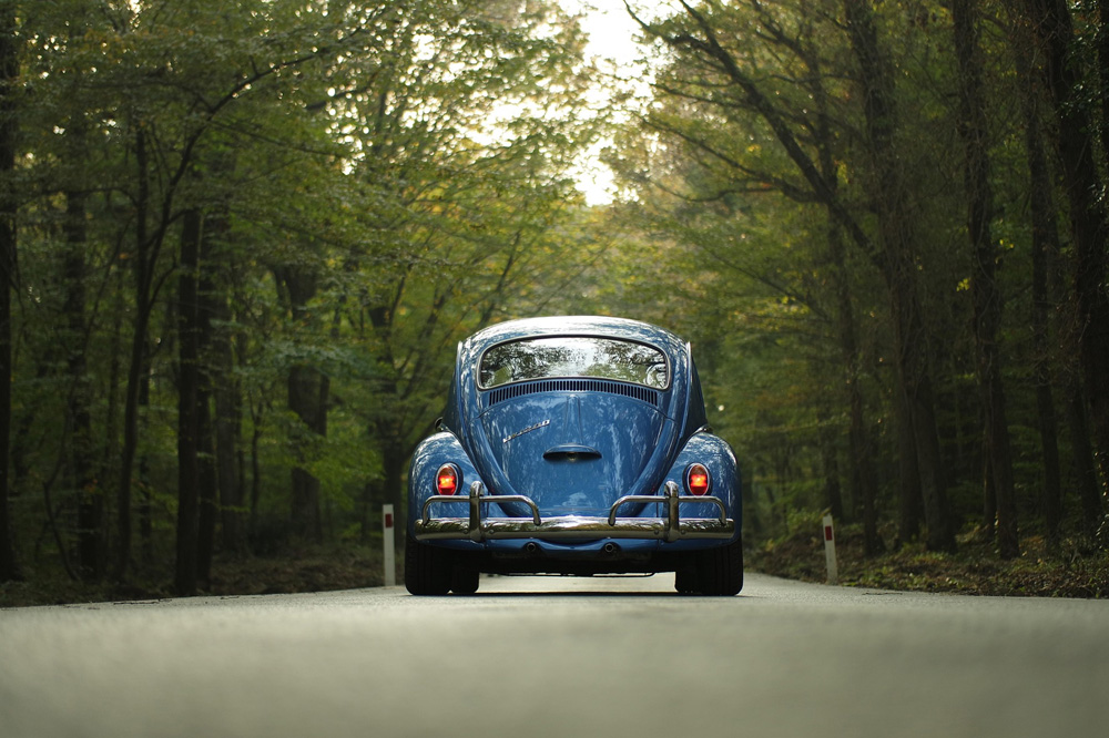 Blue Volkswagen Beetle driving through a forest road, showing classic air cooled rear engine design
