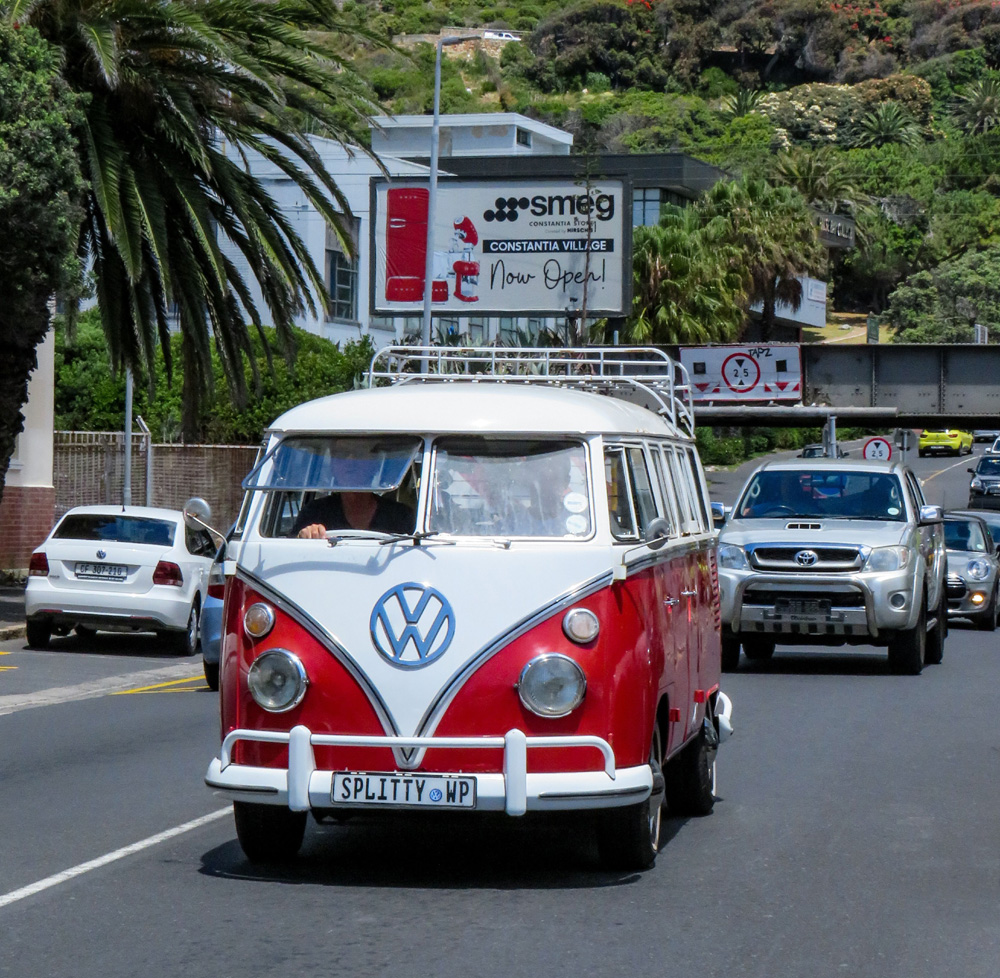 Red and white VW Type 2 driving on a sunny road with modern traffic behind it