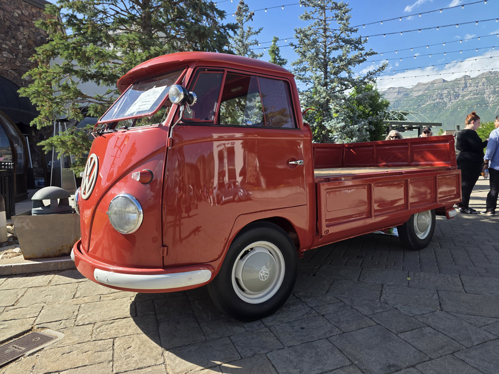 Red VW Type 2 pickup displayed outdoors with mountains and spectators nearby