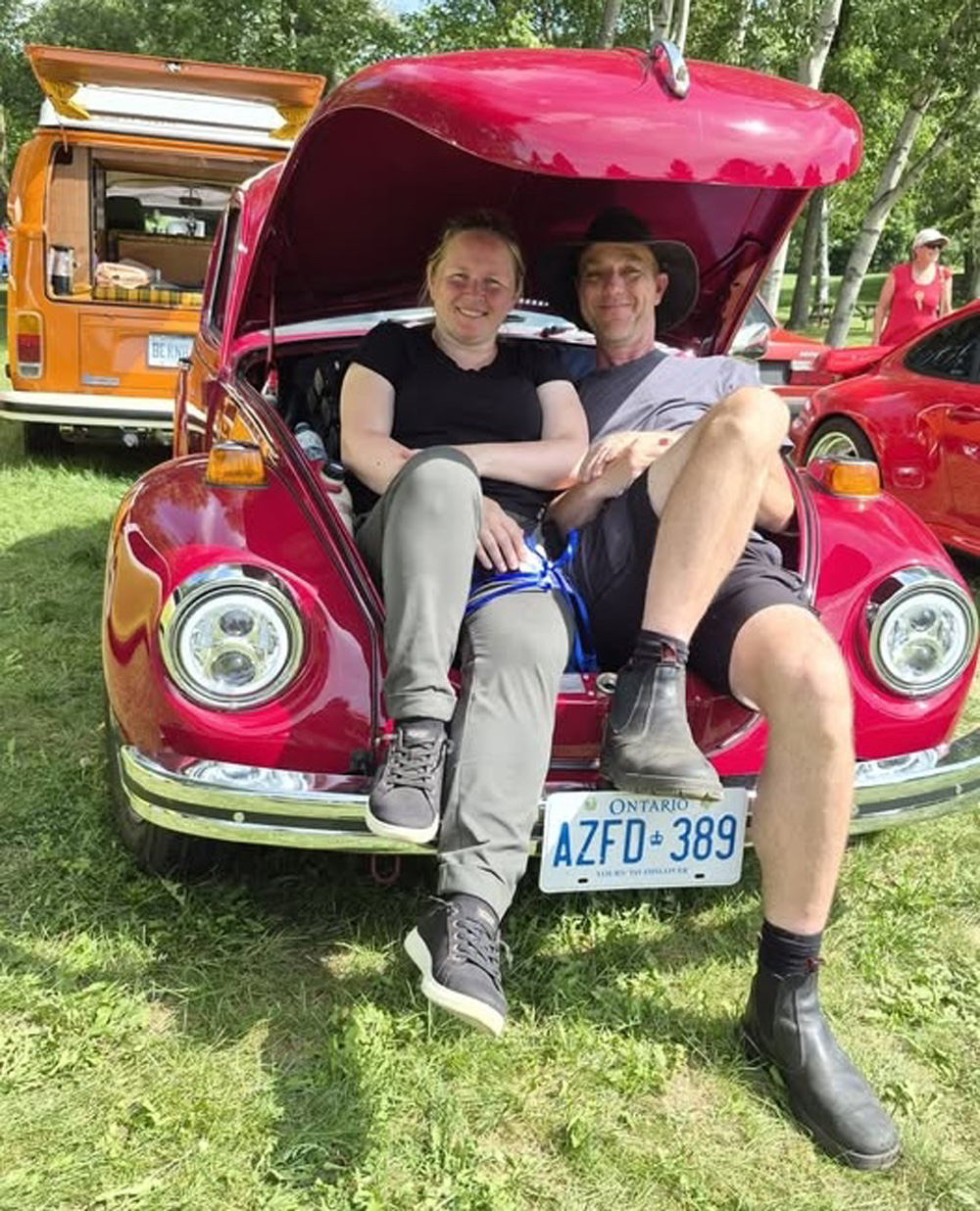 Red Volkswagen Beetle with its front trunk open at VW Legends 2026, with two people sitting inside and vintage VW campers visible behind it