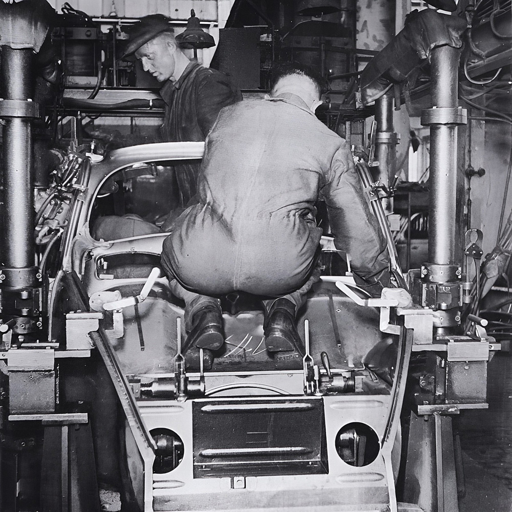 Two VW workers with the inner roof section and side panel welding jig on a Split window body.